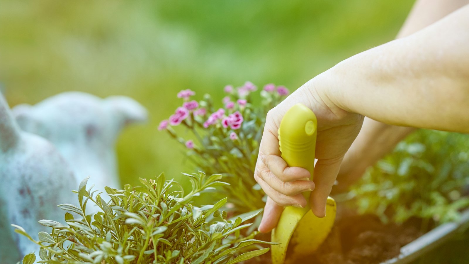 Hands carefully plant vibrant small flowers into a rectangular planter box with a yellow trowel, surrounded by lush greenery and soft natural light, highlighting spring gardening.